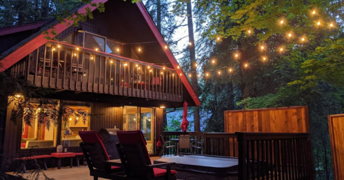 Cozy cabin surrounded by trees, featuring string lights, a deck, and outdoor seating in a serene setting.