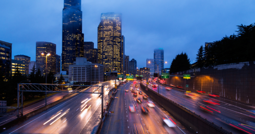 City skyline at dusk with traffic on a busy highway, illuminated buildings, and a cloudy sky.