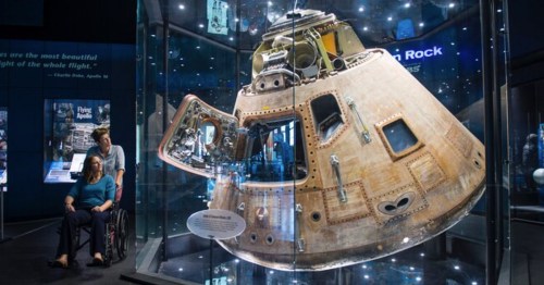 A person in a wheelchair admires a historic spacecraft displayed in a museum, surrounded by a starry backdrop.