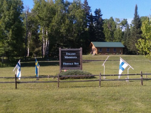 Sign for the Finland Heritage Site in Minnesota, surrounded by flags and greenery, with a cabin in the background.