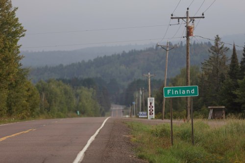 A rural road sign indicating "Finland," with trees and mountains in the background on a foggy day.