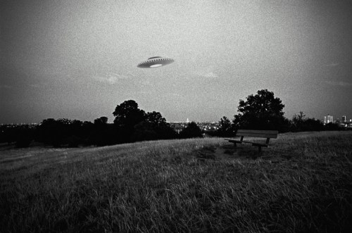 A black and white image of a UFO hovering above a grassy hill with a bench, overlooking a city skyline in the distance.