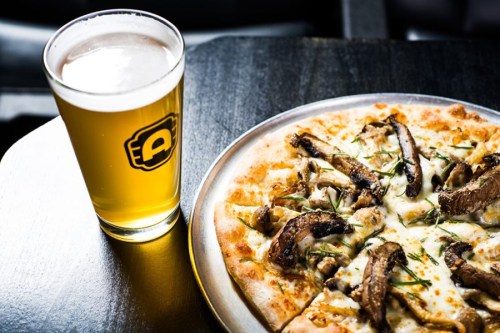 A close-up of a pizza topped with mushrooms and herbs, next to a glass of beer on a dark table.