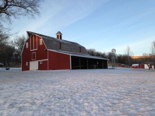 A red barn stands in a snowy landscape, with trees and a windmill in the background under a clear blue sky.