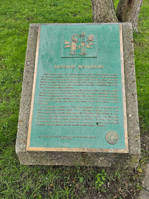 A bronze plaque titled "Mound Builders" on a stone base, set in a grassy area near trees.
