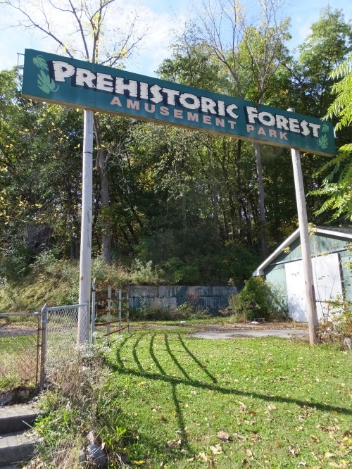 Sign for Prehistoric Forest Amusement Park, surrounded by trees and overgrown vegetation, with a building in the background.
