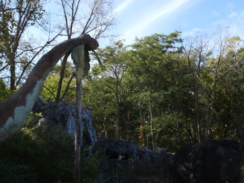 A weathered dinosaur sculpture amidst lush green trees under a blue sky.