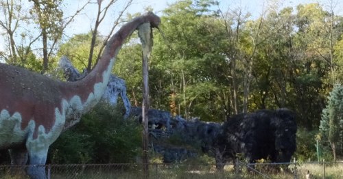 A colorful dinosaur statue stands beside a large black bison statue in a wooded area.