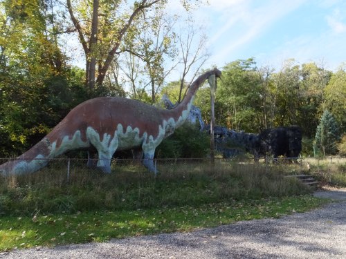 A large, colorful dinosaur statue stands in a grassy area surrounded by trees and a rocky formation in the background.