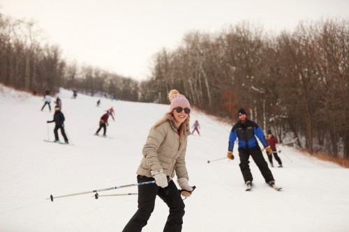 A smiling woman in a pink hat skis down a snowy slope, surrounded by other skiers in a winter landscape.
