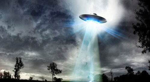 A UFO hovers in a stormy sky, emitting a bright beam of light onto a landscape with trees.
