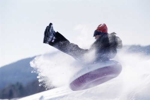 A person in winter gear joyfully sledding down a snowy hill on a tube, with snow flying around them.