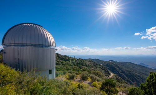 A silver observatory dome on a mountain, surrounded by greenery, under a bright sun and blue sky.