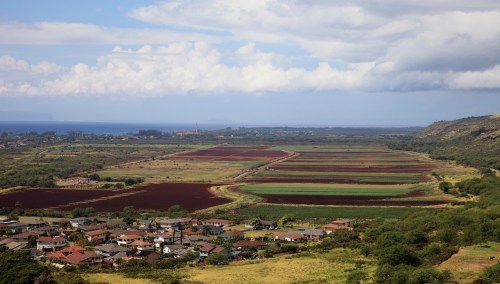 Aerial view of a rural landscape with fields, houses, and distant mountains under a partly cloudy sky.