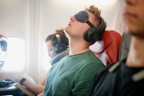 A passenger wearing headphones and a sleep mask relaxes on a plane, while another passenger reads nearby.