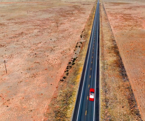 Aerial view of a long, straight road stretching through a vast, arid landscape with a red car driving along it.
