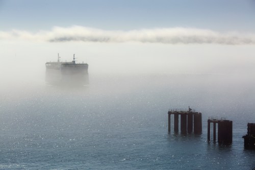 A ship emerges from fog over calm waters, with distant piers partially visible in the foreground.
