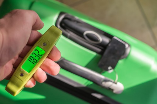 A hand holds a digital luggage scale displaying 10.3 kg above a green suitcase.