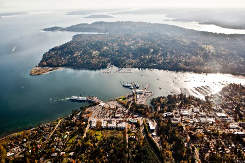 Aerial view of a coastal town with a marina, surrounded by lush greenery and water, under a clear sky.