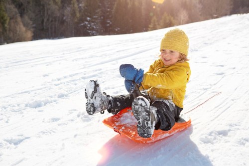 A child in a yellow jacket and hat joyfully sleds down a snowy hill on a bright winter day.
