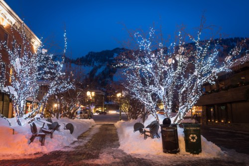 Snowy street scene at dusk, featuring trees adorned with white lights and empty chairs beside trash and recycling bins.