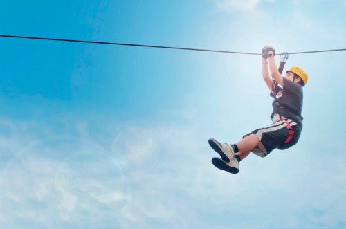 A child in a yellow helmet zip-lining against a bright blue sky with fluffy clouds.