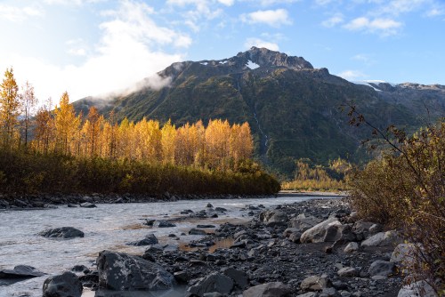 A serene river flows through a rocky landscape, framed by autumn trees and a majestic mountain under a clear sky.