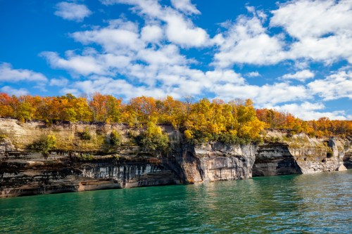 Colorful autumn trees atop a rocky cliff beside calm turquoise water under a blue sky with scattered clouds.