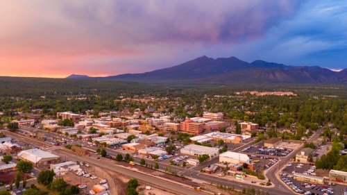 Aerial view of a town surrounded by mountains at sunset, with colorful skies and a mix of buildings and greenery.
