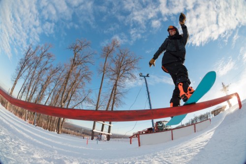 A snowboarder performs a trick on a rail in a snowy landscape with trees and a blue sky in the background.