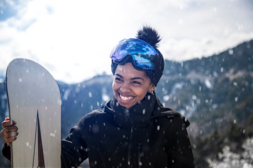 A smiling person in a black jacket and blue goggles holds a snowboard, surrounded by falling snow and mountains.