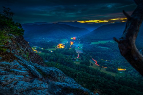 A panoramic view of a valley at dusk, with distant lights and mountains under a twilight sky.