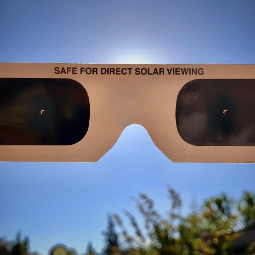 A pair of solar viewing glasses against a bright blue sky, showing small crescent shapes indicating a solar eclipse.
