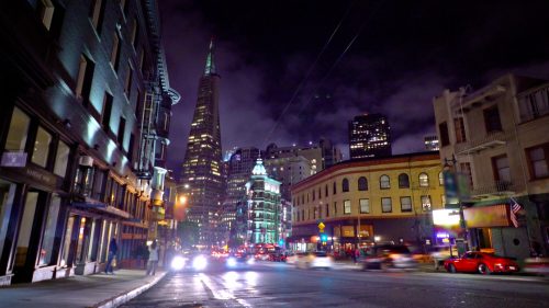 A city street at night, featuring illuminated buildings and the Transamerica Pyramid in the background.