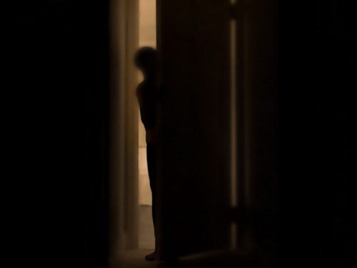 A shadowy figure stands partially behind a door, illuminated by soft light from the room beyond.