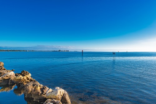 A calm blue sea under a clear sky, with rocky shoreline and distant markers visible on the water.