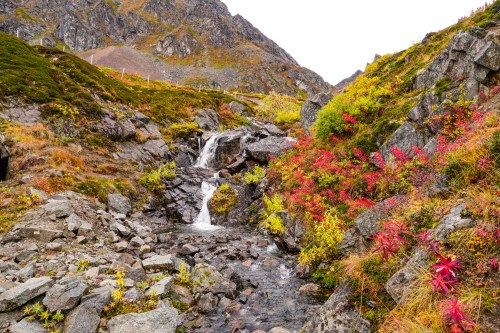 A serene mountain landscape featuring a small waterfall surrounded by colorful autumn foliage and rocky terrain.