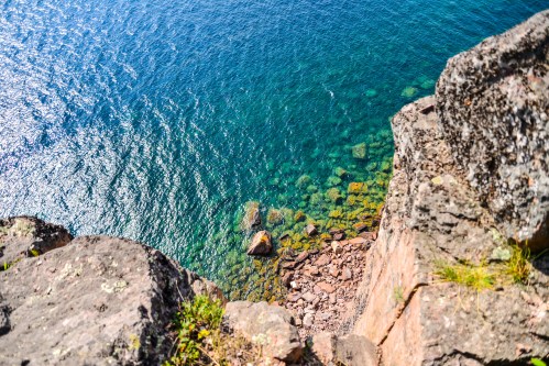 Aerial view of clear blue water and rocky shoreline from a cliff edge, with sunlight reflecting on the surface.