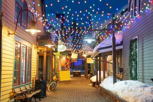 A charming alleyway adorned with colorful holiday lights and snow, featuring benches and a bicycle.