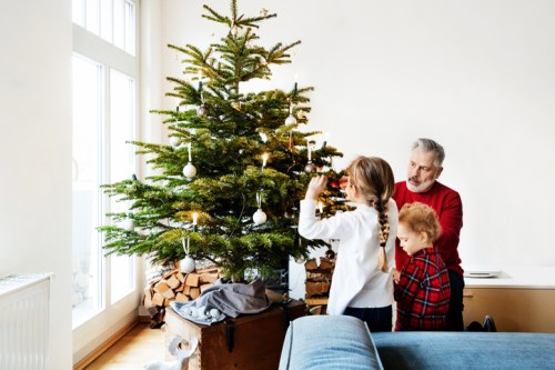 A grandfather helps two children decorate a Christmas tree in a cozy living room.