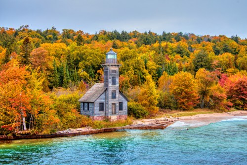 A rustic lighthouse beside a vibrant autumn forest and calm blue water, creating a picturesque coastal scene.