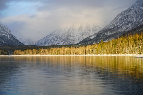 A serene lake surrounded by snow-capped mountains and trees, reflecting the landscape in calm waters.