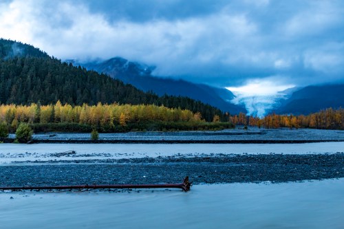 A serene landscape featuring a river, mountains, and a glacier under a cloudy sky, with autumn foliage in the foreground.