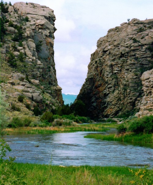 A serene river flows between towering rocky cliffs, surrounded by lush greenery and a clear blue sky.