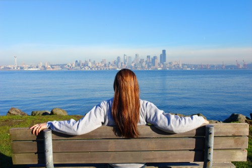 A person sits on a bench by the water, gazing at a city skyline under a clear blue sky.