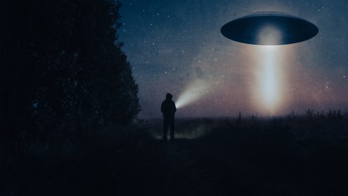 A silhouette of a person with a flashlight stands in a field, gazing at a hovering UFO emitting a beam of light.