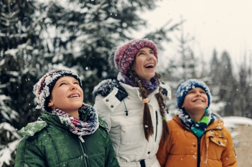 A woman and two children smile and look up at falling snow in a snowy forest. They are bundled in winter clothing.