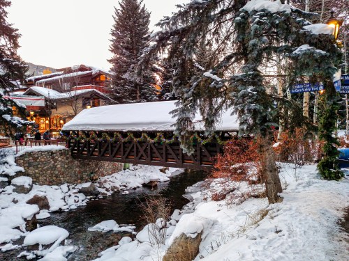 A snow-covered bridge adorned with holiday decorations spans a stream, surrounded by trees and festive buildings.