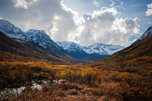 A scenic mountain valley with snow-capped peaks, colorful autumn foliage, and a clear blue sky with clouds.