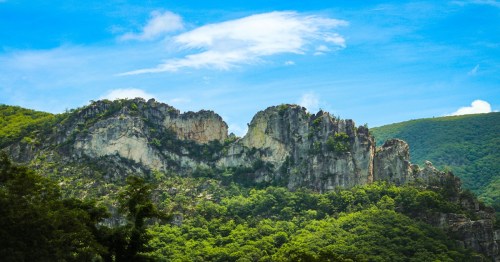 A rugged mountain peak rises above lush green hills under a bright blue sky with scattered clouds.